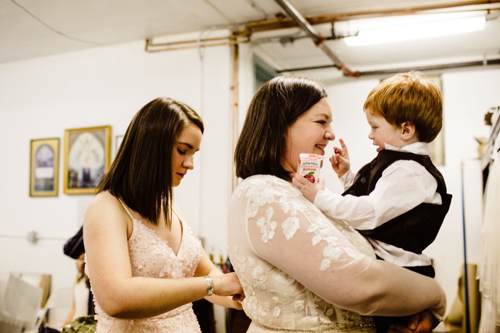bride holds her infant son in her arms as she puts on wedding dress