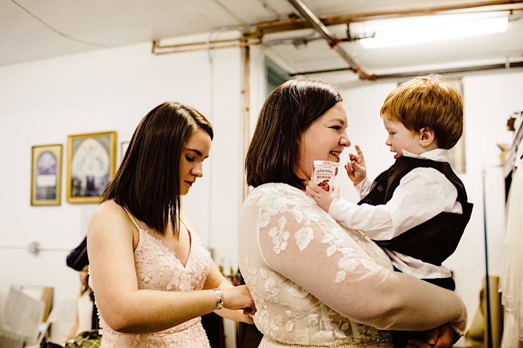 smiling bride holds son while getting her dress on